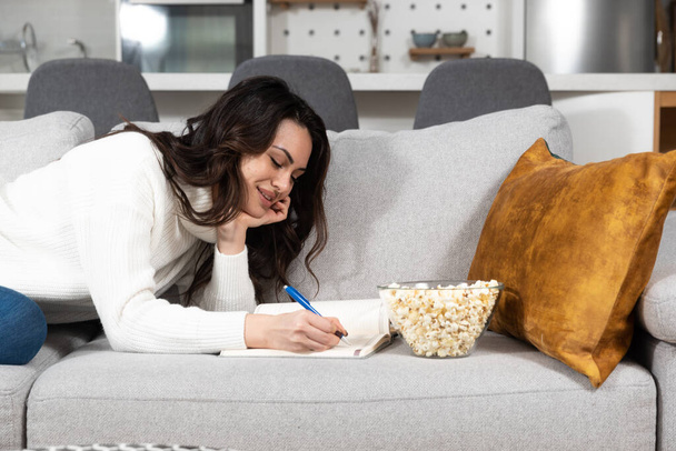 Young beautiful girl writing a love letter to her boyfriend or writing in the diary while she is smiling and sitting on the sofa in her room. Successful female having business ideas. - Foto, afbeelding