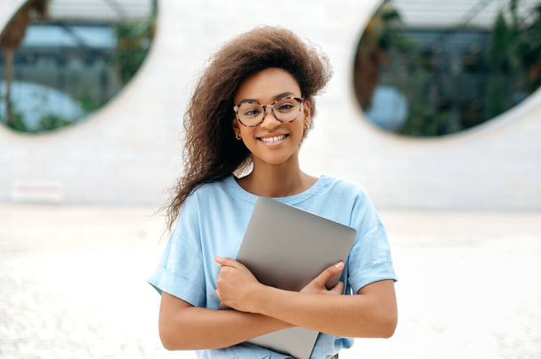 Beautiful friendly positive curly haired african american young woman with glasses, in a blue shirt, freelancer, student, businesswoman, holding laptop, standing outdoors, looking at camera, smiling - Fotó, kép