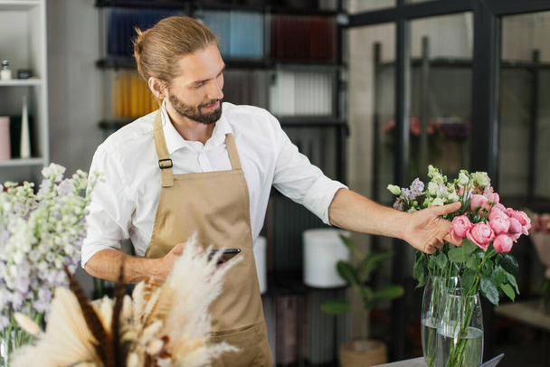 Man florist at her own floral shop taking care of flowers. People, business, sale and floristry concept. Happy smiling male florist making different flower bunch at flower shop. - 写真・画像