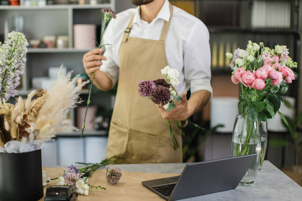 Attractive bearded male gardener in the flower shop make bouquet for a holiday. Family flowers business. Lifestyle flower shop. Beautiful flower composition. Detail. Close up. - Фото, зображення