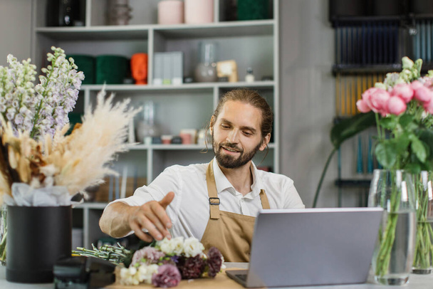 Portrait of attractive man florist working on computer at workplace. Concentrated bearded male sitting at counter in floral store and typing on laptop. - Fotografie, Obrázek