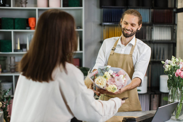 Attractive young woman shopping and buying bouquet in bearded male florist in flower shop. Female customer paying with card for her purchase bouquet of flowers. - Valokuva, kuva