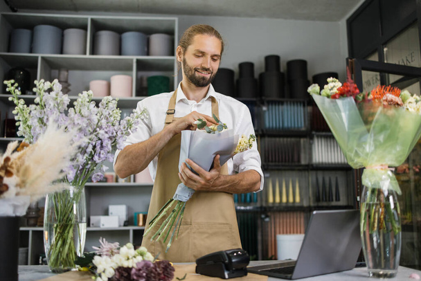 Portrait of male florist in apron working in flower shop composing bouquets with fresh flowers for sale, floris making workshop for young employee during time in greenery. - Photo, image