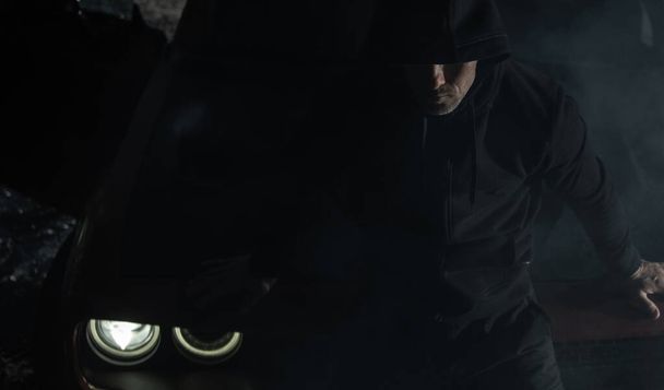 Valkoihoinen mies mustissa vaatteissa istuu muskeliautonsa konepellillä pimeydessä. Huumekauppias pitää silmällä hänen aluettaan öisin. Rikollisen maailman teema. - Valokuva, kuva