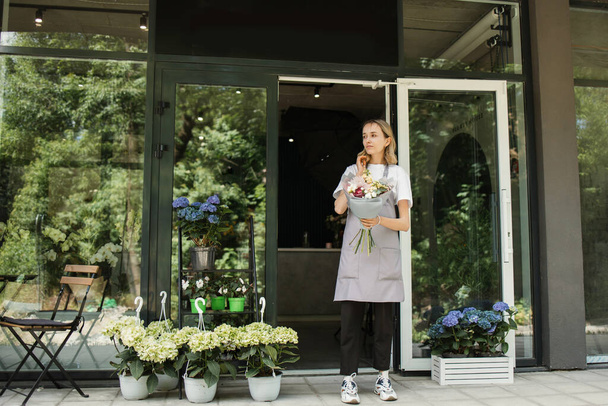 Business Woman near flower shop door. Photo of beautiful woman standing in front of flower shop. Female Florist Outside Shop. Woman working at flower shop smiling - Valokuva, kuva