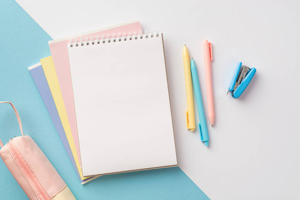 Концепція шкільного приладдя. Фотографія з видом зверху барвистого канцелярського відкритого ноутбука ручки пенал і міні степлер на біколорному синьо-білому тлі з порожнім простором
 - Фото, зображення