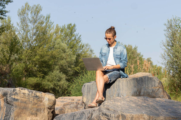 Na volné noze pracující mladý muž se svým notebookem z přírody sedí na kameni mezi stromy lesa v létě - Fotografie, Obrázek