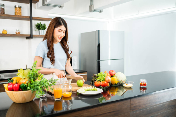 A happy young Asian woman preparing a healthy salad with vegetables on a cutting board on the home kitchen counter. Food cooking for couple husband and wife in a good relationship. - Foto, Imagen