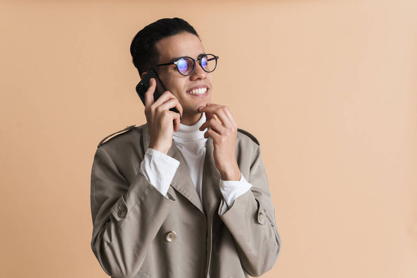 Young middle eastern man in eyeglasses talking on mobile phone isolated over beige background - 写真・画像