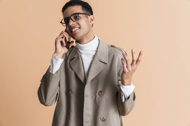 Young middle eastern man gesturing while talking on mobile phone isolated over beige background - Fotó, kép