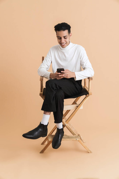 Young middle eastern man using cellphone while sitting on chair isolated over beige background - Foto, Bild