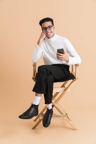 Young middle eastern man using cellphone while sitting on chair isolated over beige background - Foto, immagini