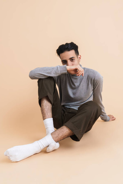 Young middle eastern man looking at camera while sitting on floor isolated over beige background - Fotografie, Obrázek