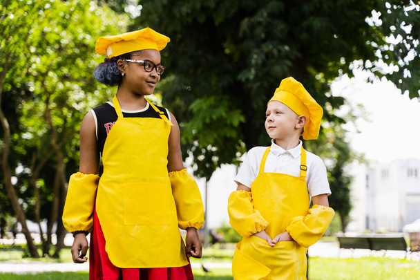 Communication between multiracial friends of black african and caucasian cook children in yellow chefs hat and apron discussing and having fun together - Foto, Imagem