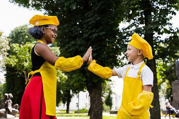 Black african and caucasian cook child in yellow chefs hat and apron clap each others hands and give five. Black african sister cooking and caucasian brother smiling and having fun together - Foto, Imagen