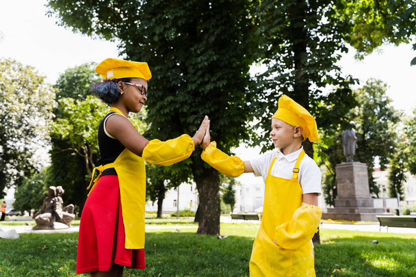 Black african and caucasian cook child in yellow chefs hat and apron clap each others hands and give five. Black african sister cooking and caucasian brother smiling and having fun together - Фото, изображение