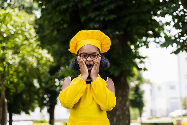 Шокована чорношкіра африканська дитина готує дівчину в капелюсі шеф-кухарів і жовтий фартух у формі тримає щоки і дивує. Творча реклама для кафе чи ресторану
 - Фото, зображення