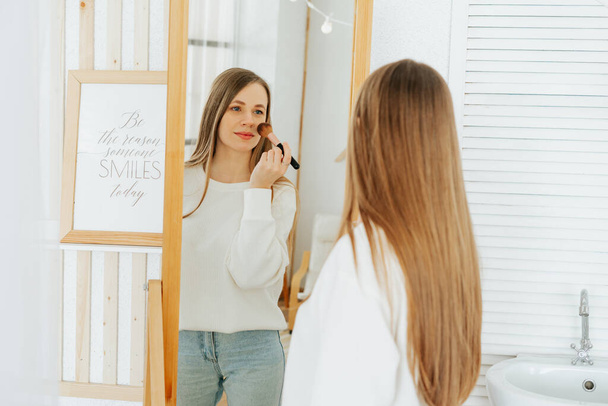 Hermosa joven mujer caucásica sonriente con el pelo largo de pie cerca del espejo, haciendo maquillaje y mirando el reflejo en la sala de estar en casa. - Foto, imagen