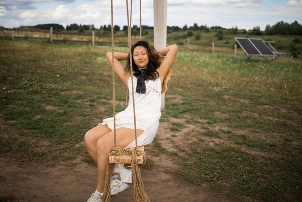Cheerful asian lady in white dress leaning back in her swing in sunny golden countryside. - Foto, imagen
