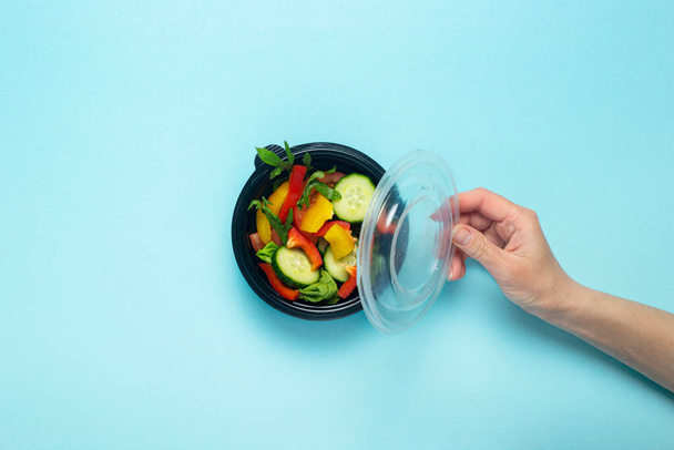 A female hand holds an open plastic disposable plate with vegetable salad on a blue background. Top view, flat lay. - Foto, afbeelding