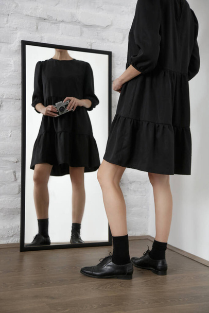 Junge Frau in schwarzem Kleid und Oxford-Schuhen hält Retro-Kamera vor Spiegel.  - Foto, Bild