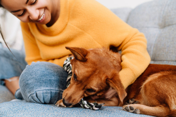 Young hispanic woman smiling and petting her dog while resting on couch at home - Photo, Image