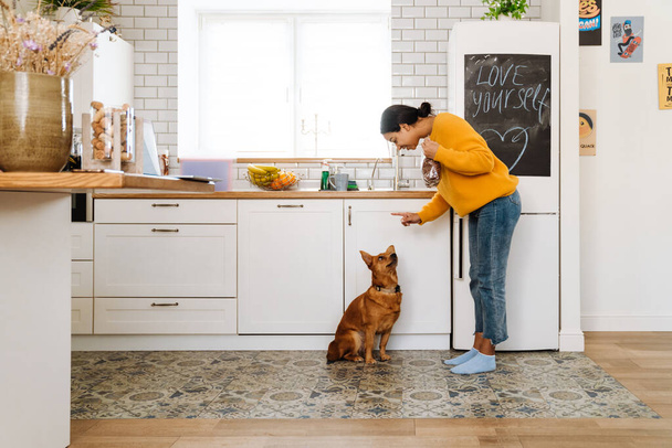 Young hispanic woman smiling while feeding her dog in kitchen at home - Fotografie, Obrázek
