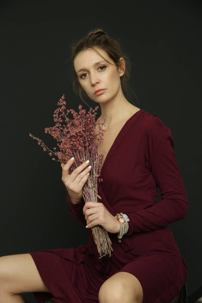 Serie van studio foto 's van jonge vrouwelijke model in Bourgogne wrap jurk met een boeket van gedroogde rode bloemen - Foto, afbeelding