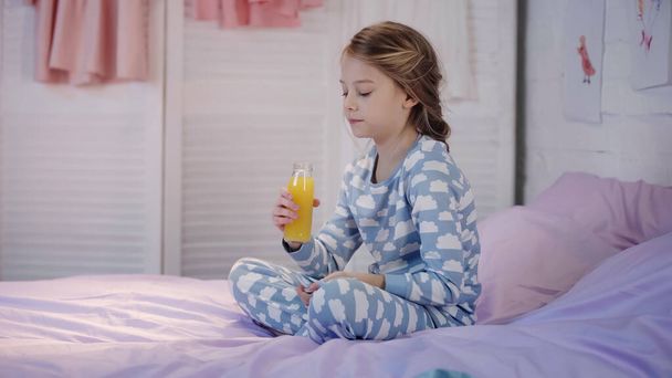 Girl in pajama holding bottle of orange juice on bed in evening  - Φωτογραφία, εικόνα