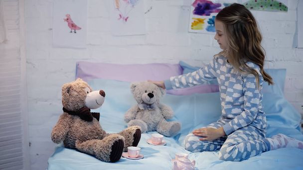 Side view of child in pajama touching sot toy near tea cups on bed at home  - Foto, Imagen