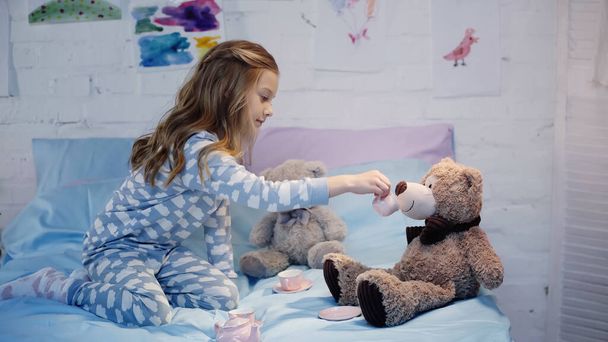 Side view of girl in pajama holding cup near teddy bear on bed  - Foto, Imagen