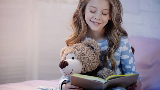 Neşeli çocuk oyuncak ayıyı tutuyor ve yatakta kitap okuyor.  - Fotoğraf, Görsel