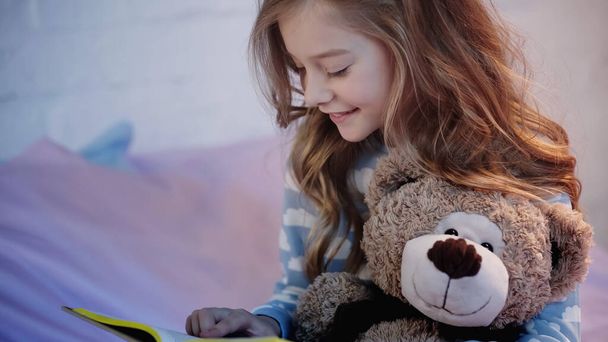 Cheerful preteen child in pajama reading book near soft toy in blurred bedroom  - Zdjęcie, obraz