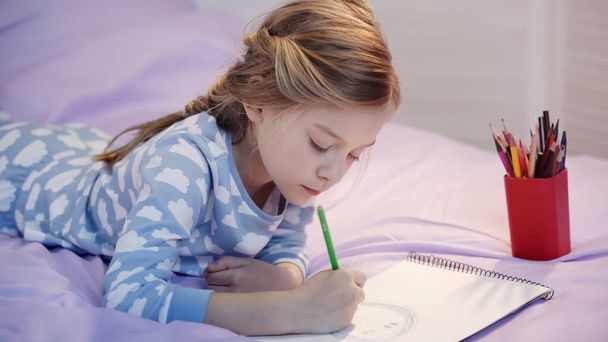 Preteen kid in pajama drawing on sketchbook on bed  - 写真・画像