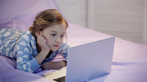 Preteen girl in pajama looking at laptop on bed in evening  - Φωτογραφία, εικόνα