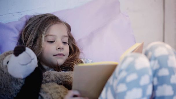 Preteen kid reading book while lying near teddy bear on bed  - Foto, imagen