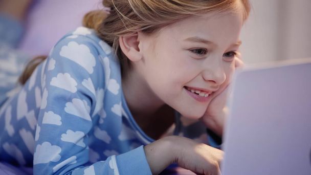 Positive preteen kid in pajama looking at blurred laptop on bed  - Фото, изображение