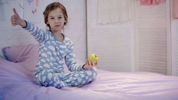 Dítě v pyžamu ukazuje a drží jablko na posteli ve večerních hodinách  - Fotografie, Obrázek