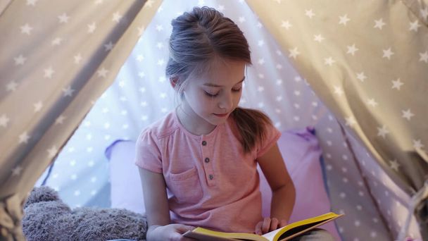 Preteen child in t-shirt reading book in wigwam in evening  - Photo, Image