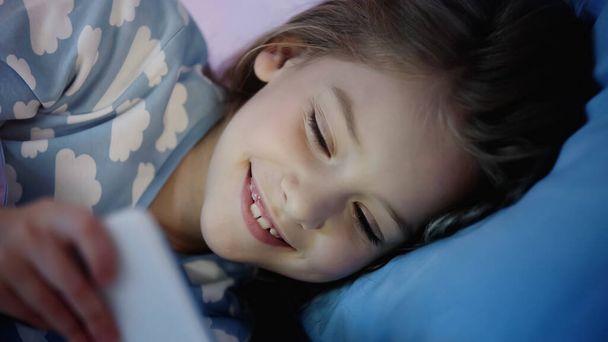 Cheerful kid in pajama looking at smartphone on bed in evening  - Fotografie, Obrázek