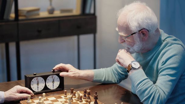 bearded man fixing time on chess clock while gaming with senior friend at home - Foto, Bild