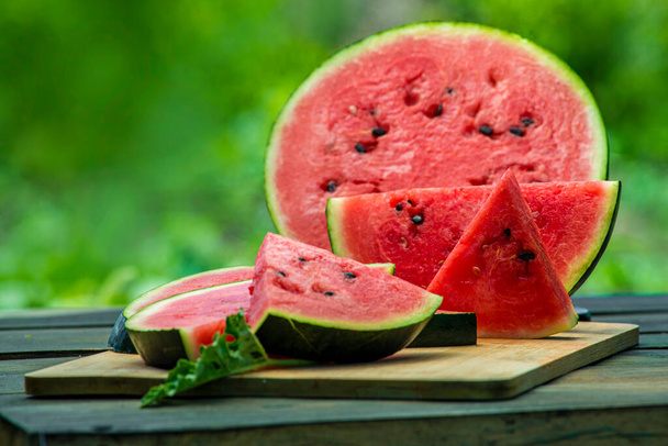 Delicious watermelon  outdoors wooden  table, GREEN  natural background  Slice sunny day, Heap,  Fresh ripe red, leaf, summer sunny garden juice drink organic sweet fruit, vegan food Tasty sliced board plate - Фото, изображение