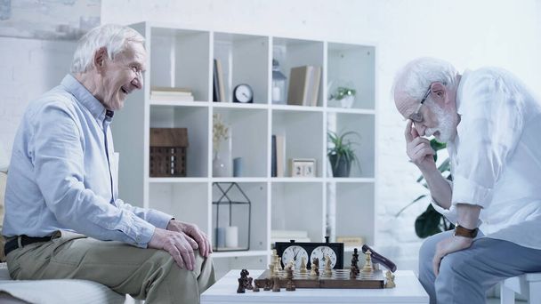 side view of senior man thinking near chessboard and laughing friend  - Фото, изображение