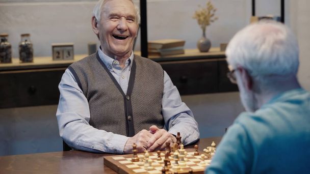 excited elderly man laughing near chessboard and blurred friend  - Foto, Imagen