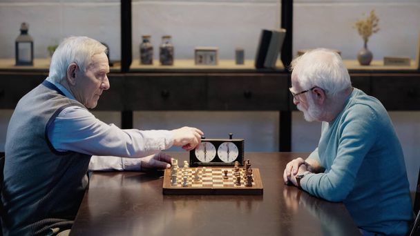side view of senior man fixing time on chess clock near friend in living room - 写真・画像