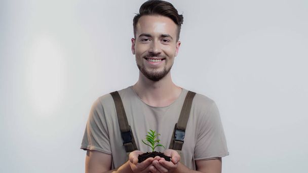 Smiling gardener holding soil and plant on grey background - Фото, изображение