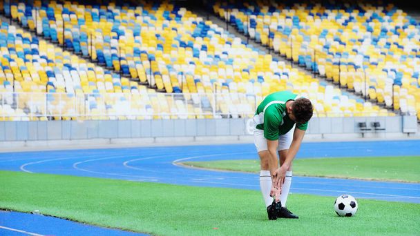 young football player in uniform stretching near ball of green field  - Valokuva, kuva