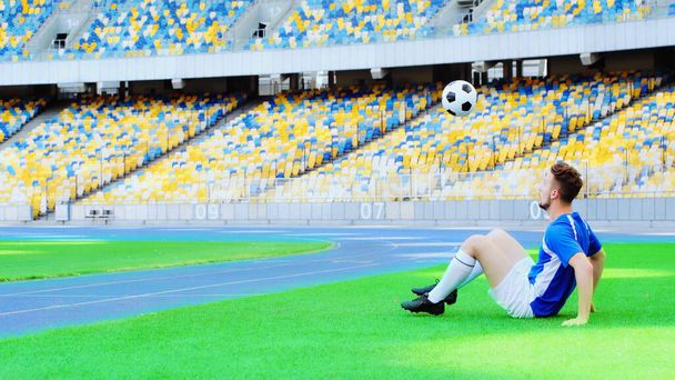 football player sitting on green grass and bouncing ball with legs - Foto, imagen