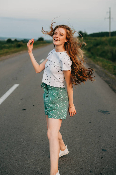 Ginger woman walking away from the camera down a country road in late evening, fields on either side. Summer vacation. Healthy lifestyle. Modern lifestyle. - 写真・画像