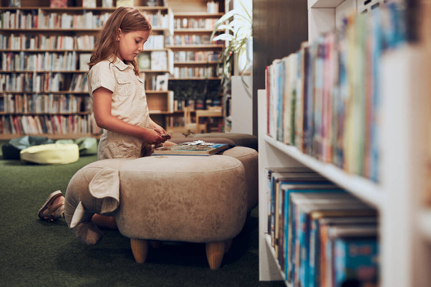 Schoolgirl doing puzzle and reading book in school library. Primary school pupil is involved in book with jigsaw. Smart girl learning from book. Benefits of everyday reading. Child curiosity - Fotografie, Obrázek